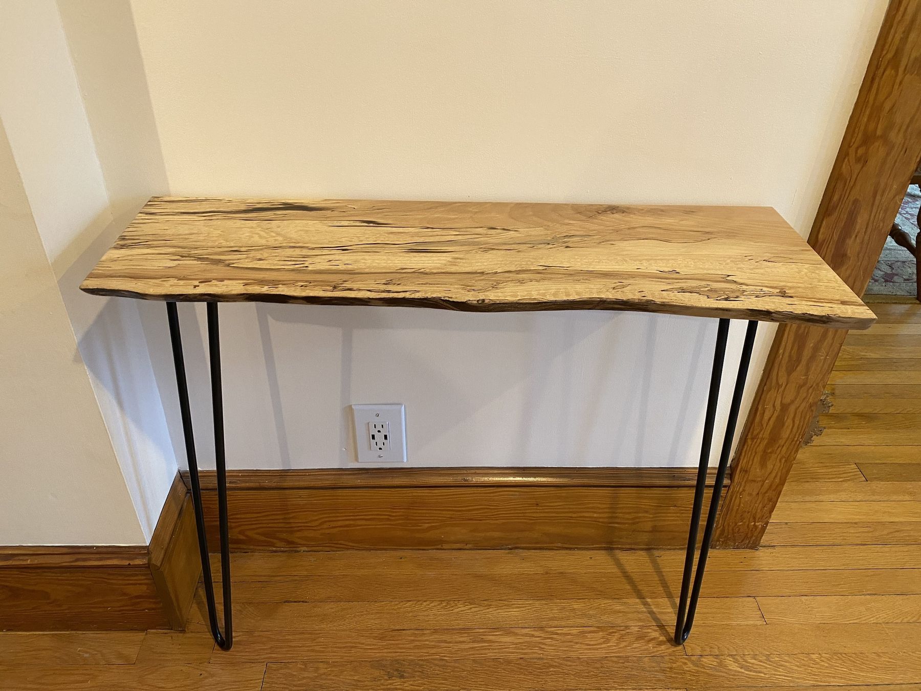 A narrow live-edge console with dark spalting lines through a pale slab, standing on black steel hairpin legs against a cream wall.